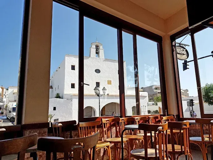 Vista interior de una cafetería con un gran ventanal que da a un edificio blanco con campanario en un día soleado. Sillas y mesas de madera en el interior.
