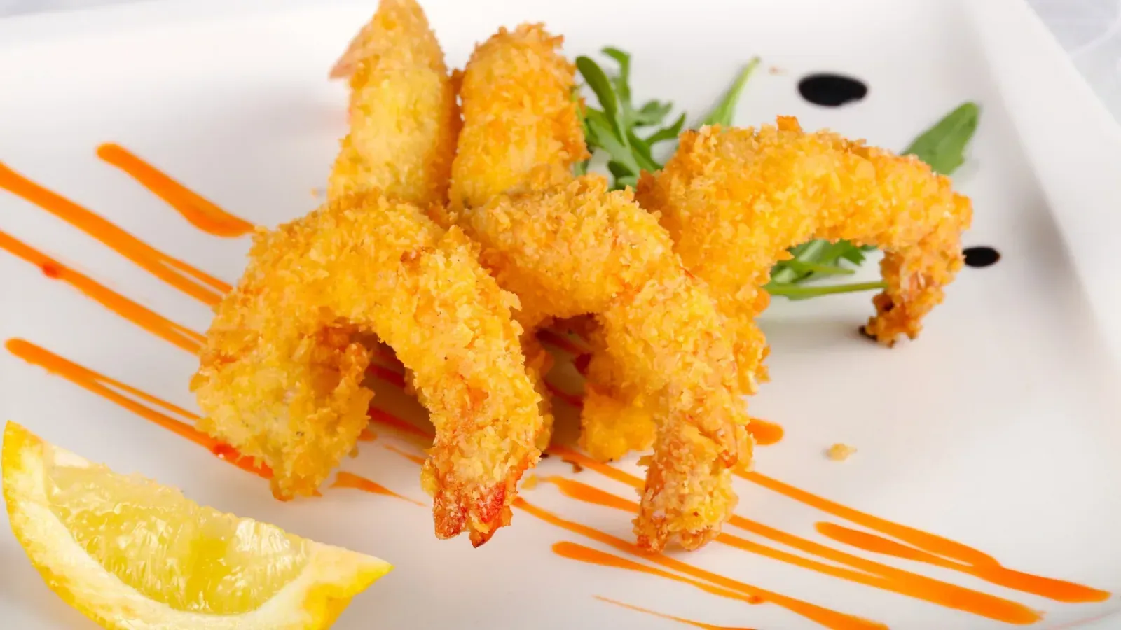 Camarones fritos en un plato blanco con un chorrito de salsa de naranja, rodaja de limón y rúcula.
