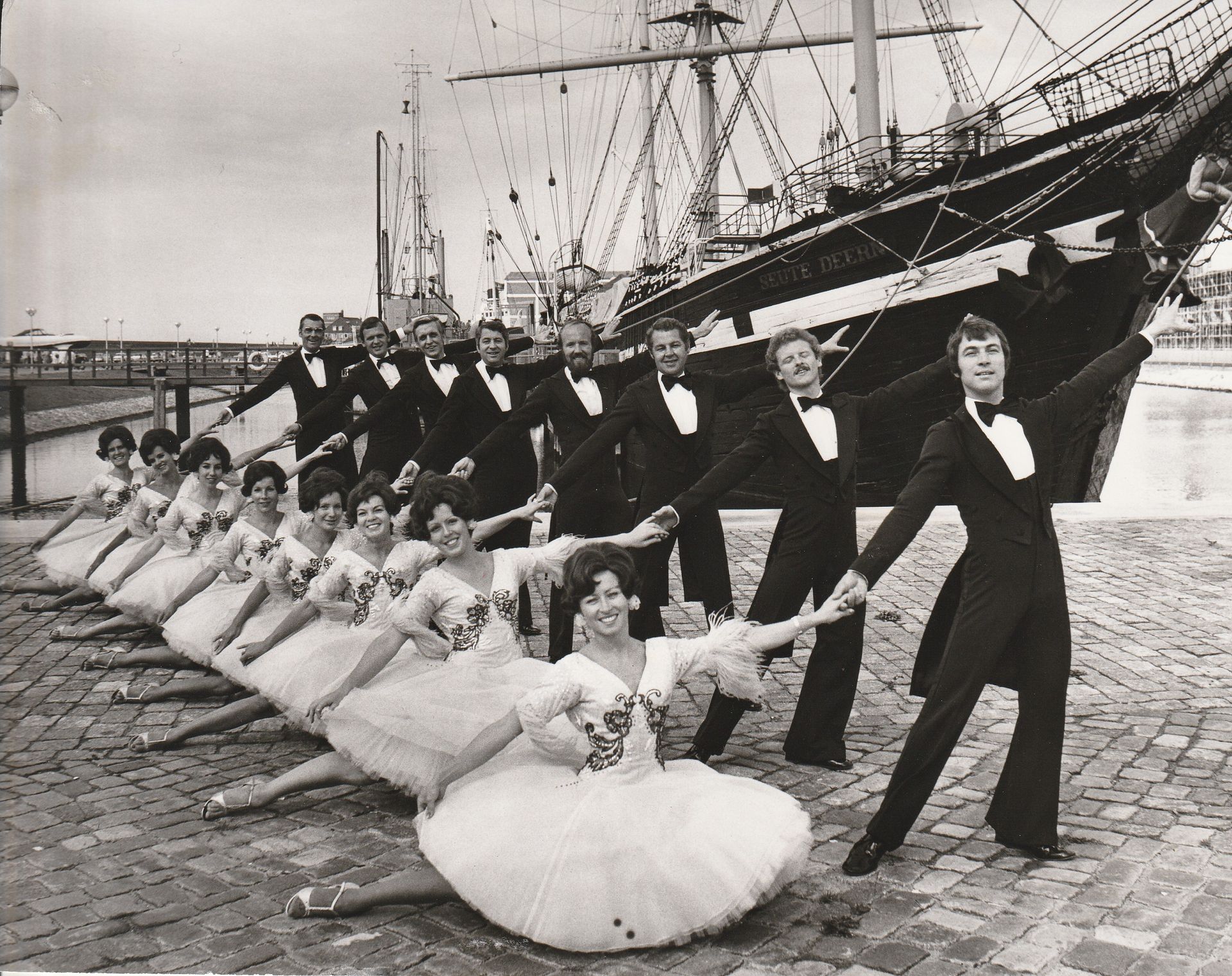 Ein Schwarzweißfoto einer Gruppe tanzender Menschen vor einem Schiff