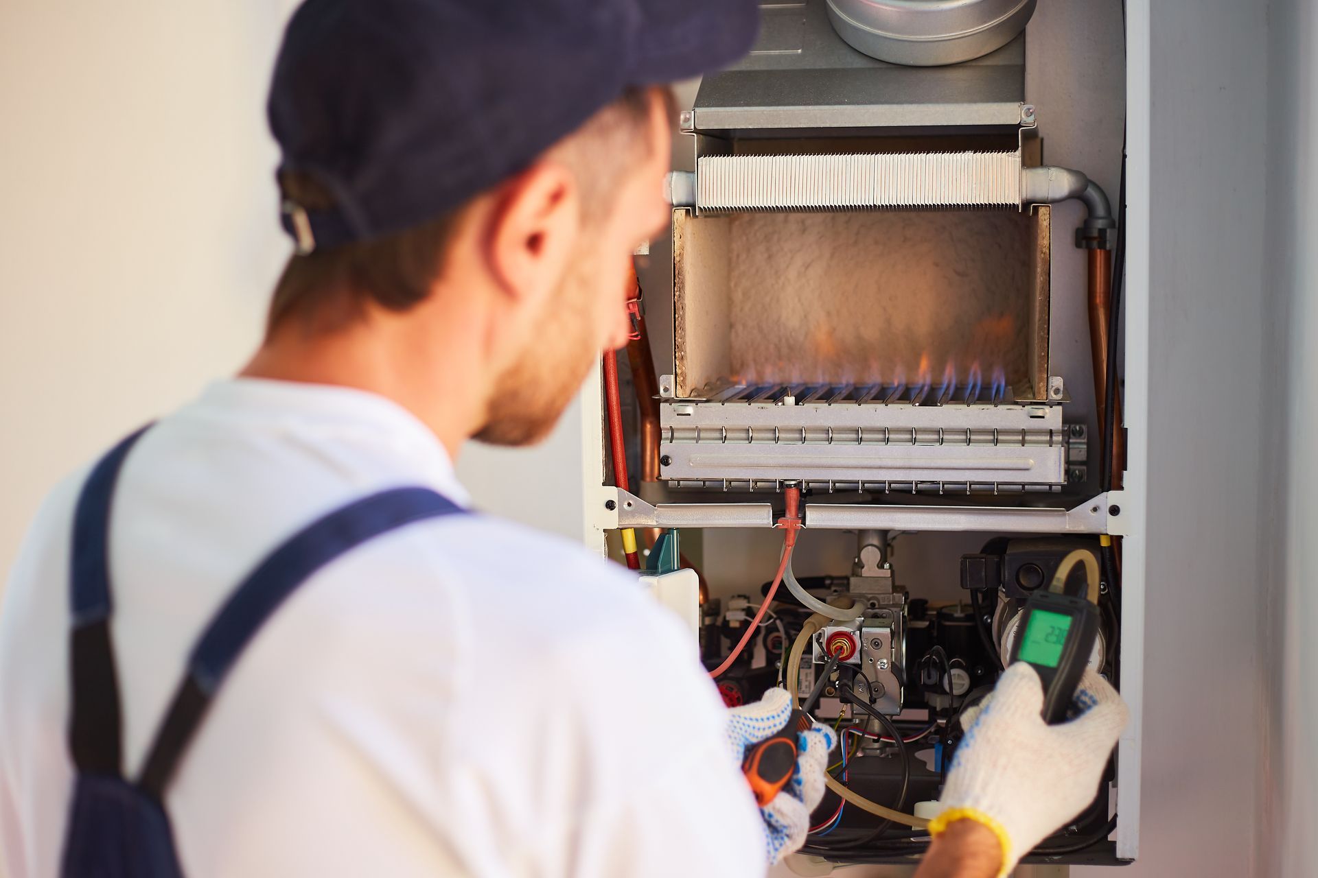 Un technicien inspecte une chaudière. Des flammes sont visibles à l'intérieur. Le technicien porte une casquette, une combinaison et des gants, et tient un appareil de mesure.