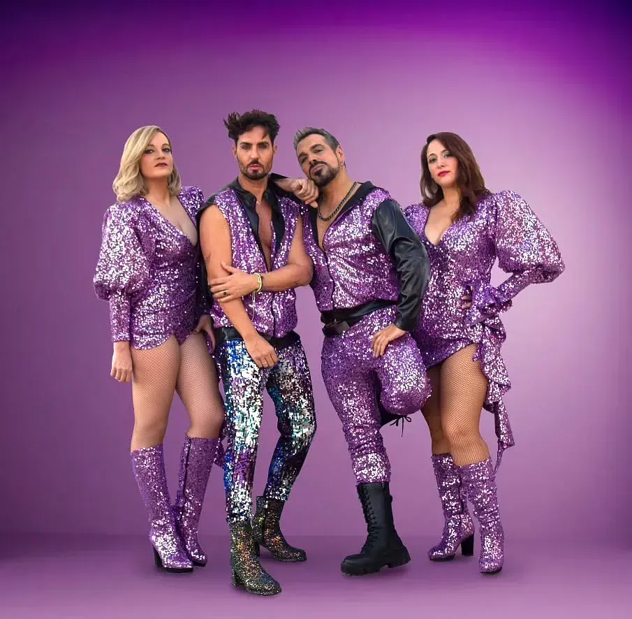 Cuatro personas con brillantes atuendos morados posando sobre un fondo morado. Dos mujeres y dos hombres.