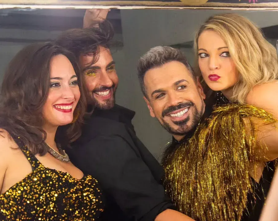 Cuatro personas sonriendo y posando para una selfi frente a un espejo. Dos mujeres, dos hombres. Brillantina, oro.