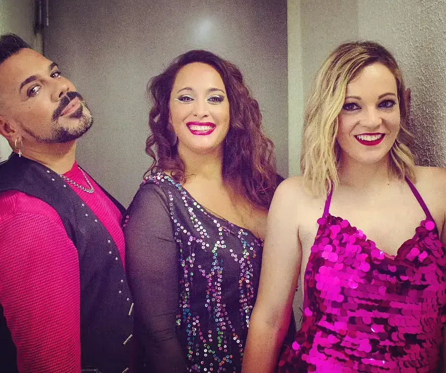 Tres personas sonriendo, posando juntas en el interior: un hombre de rosa y dos mujeres con vestidos brillantes.