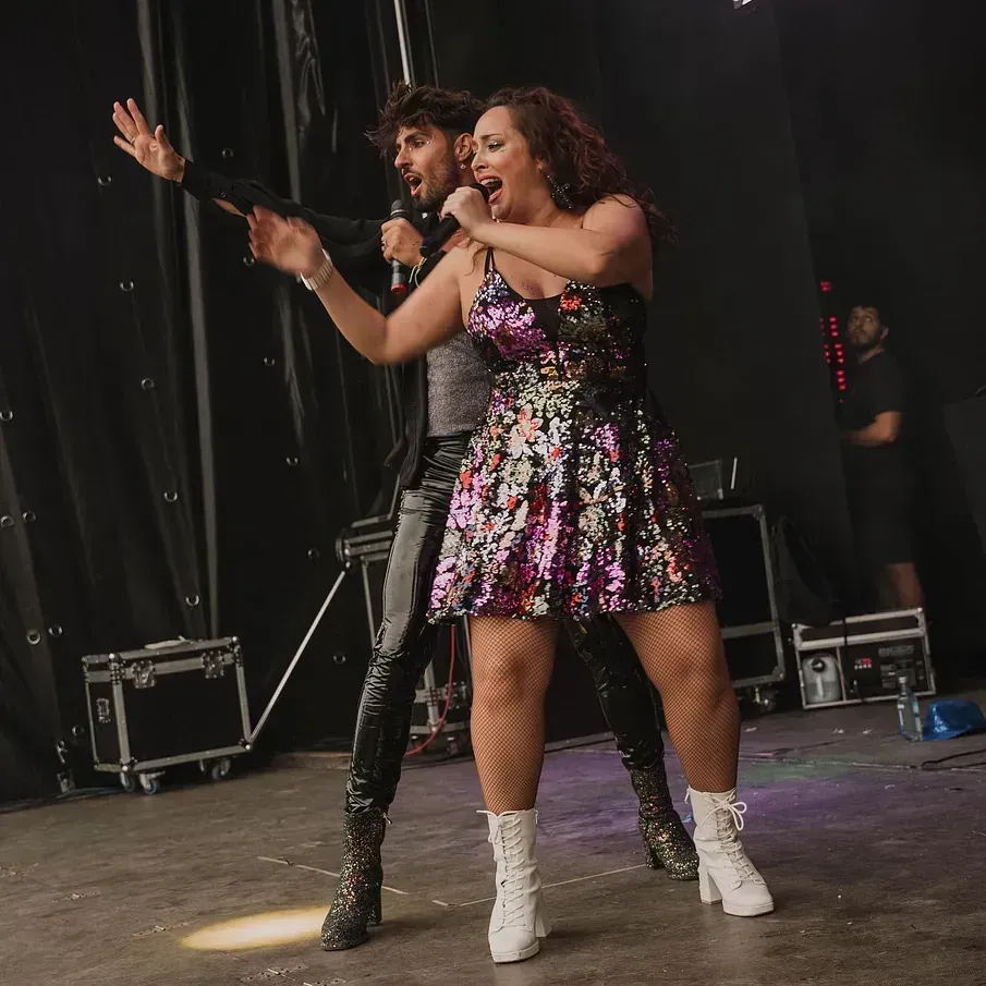 Un hombre y una mujer cantan en el escenario. Una mujer con un vestido de lentejuelas y un hombre con un traje negro, ambos con micrófonos.