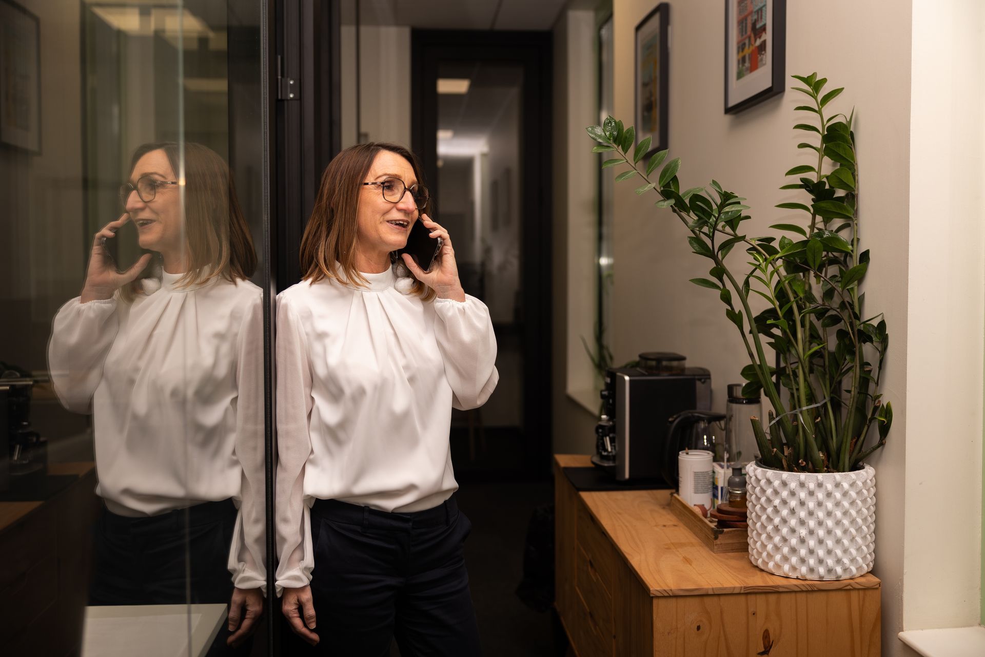 Une personne en chemisier blanc parle au téléphone portable, debout dans un couloir à côté d'une grande plante en pot.