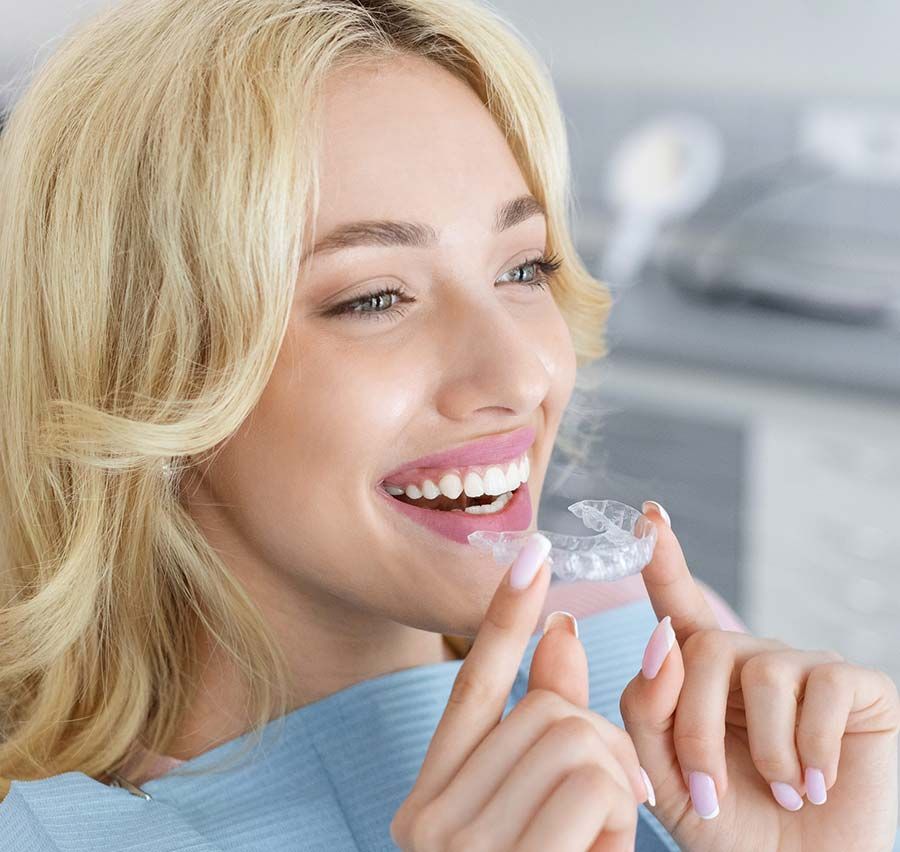 Mujer sonriente sosteniendo un alineador dental transparente cerca de su boca en un entorno clínico luminoso.