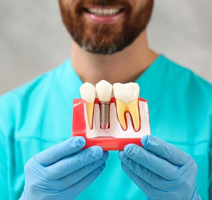 Dentista sosteniendo un modelo de dientes con implante, encías y coronas, con guantes azules y bata quirúrgica.