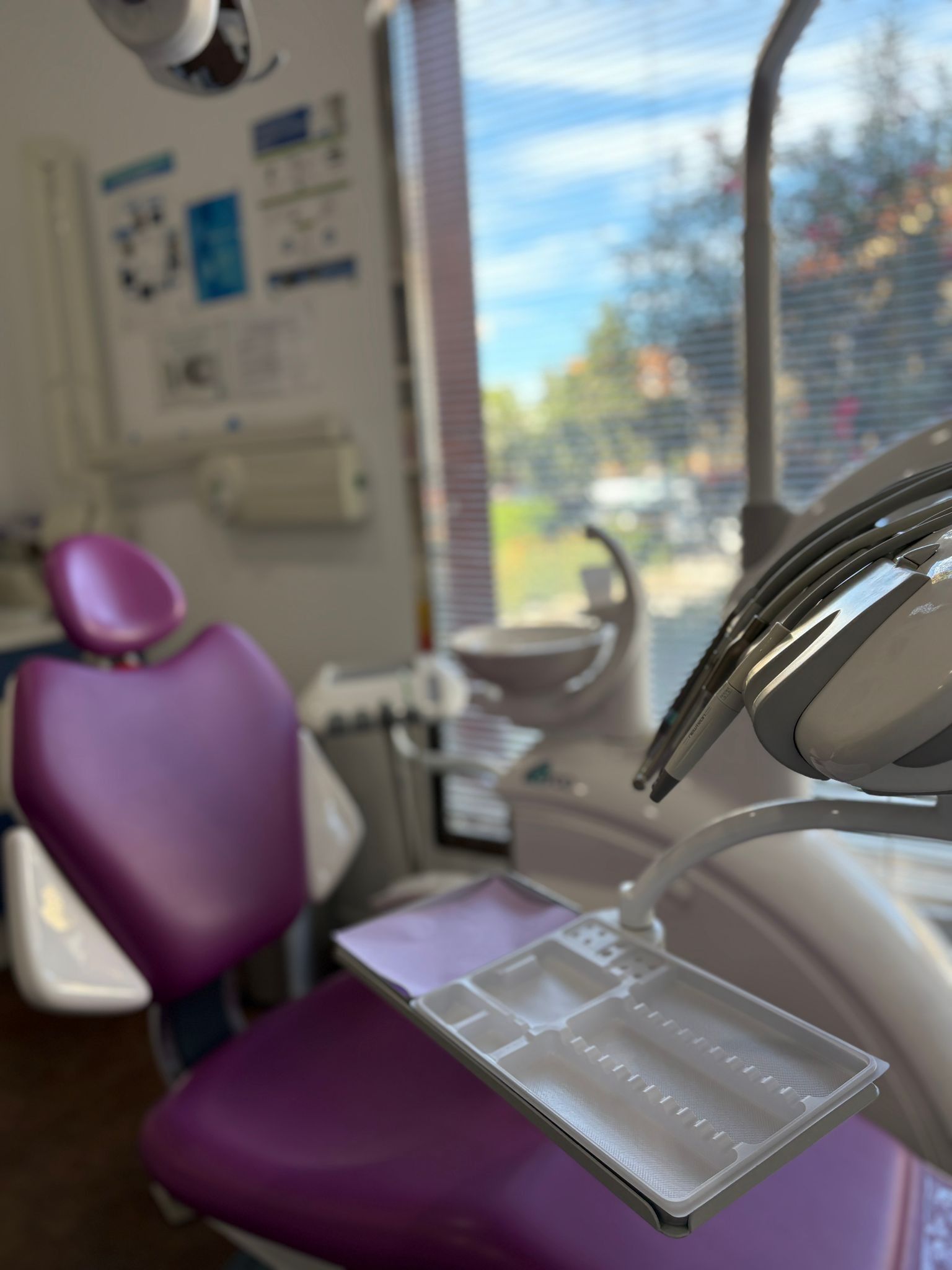 Sillón dental morado con bandeja en un consultorio dental muy iluminado, vista desde una ventana.