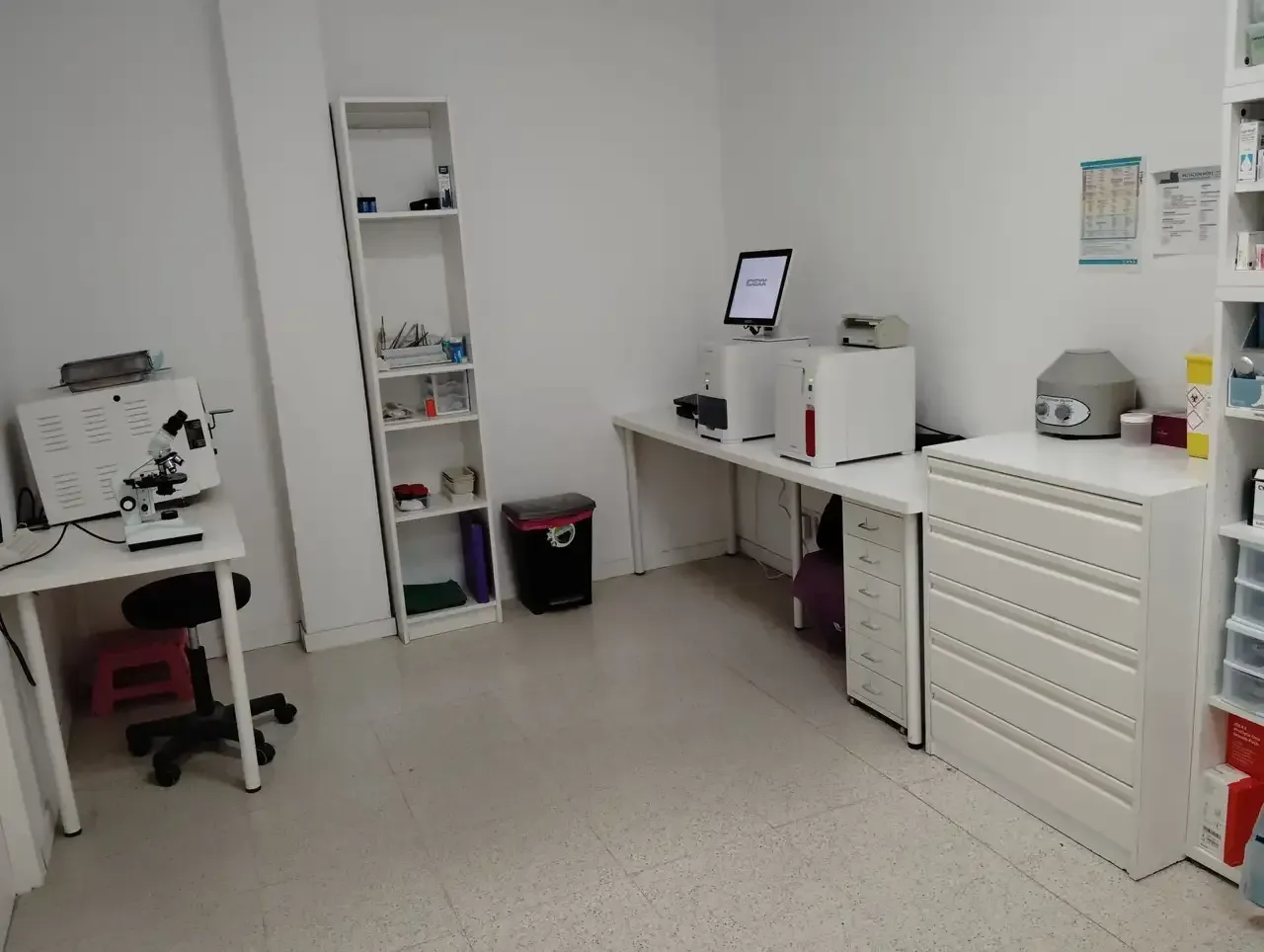 Una pequeña habitación con una mesa, una impresora y una computadora. Otra mesa contiene un proyector y una canasta.