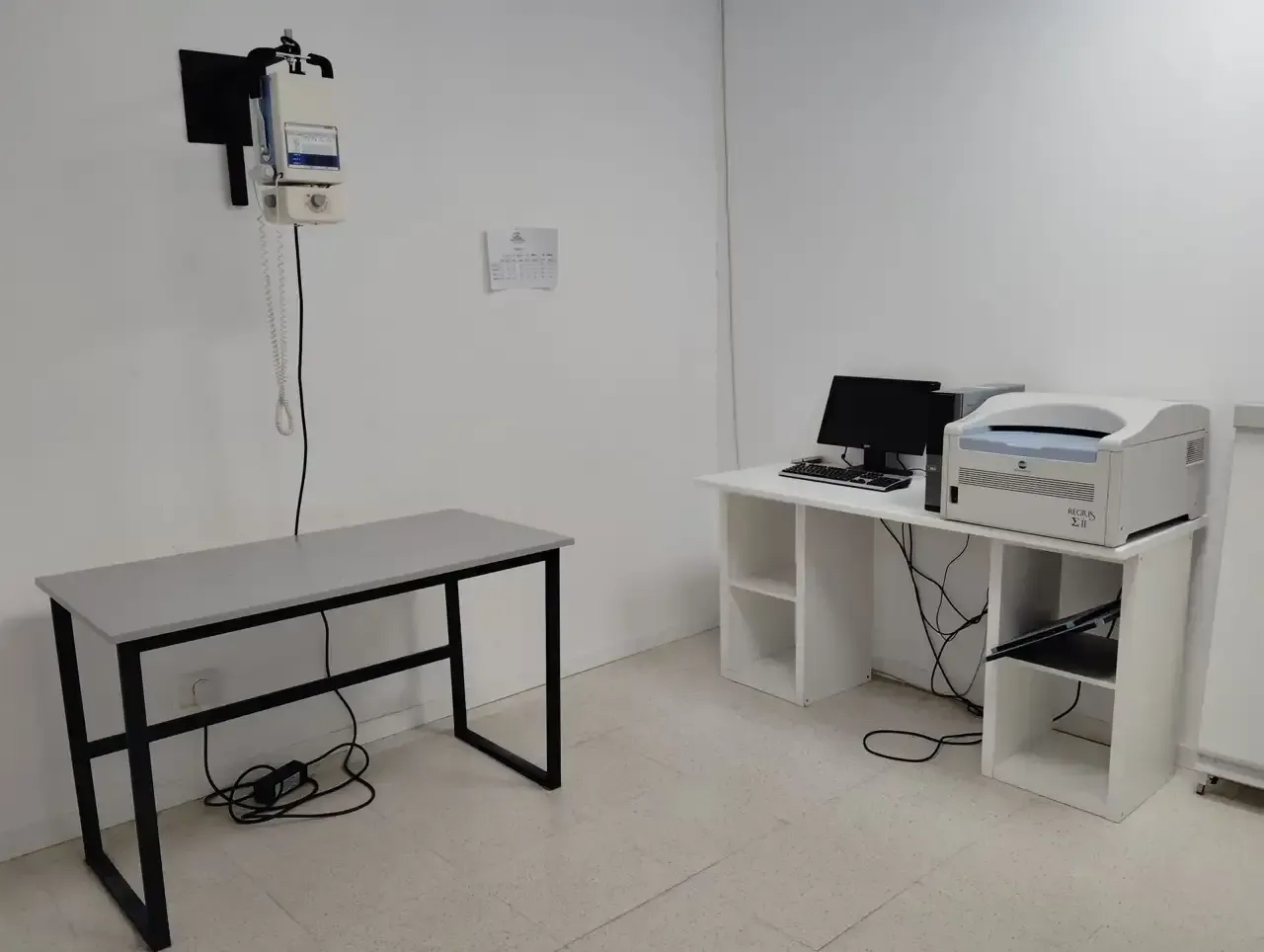 Una pequeña habitación con una mesa, una impresora y una computadora. Otra mesa contiene un proyector y una canasta.