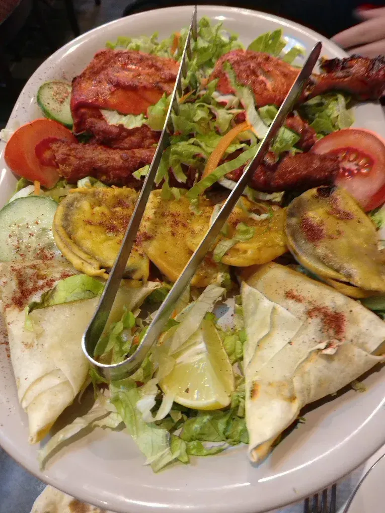 Un assortiment de mets indiens dans une assiette : poulet tandoori, légumes grillés, samosas et salade verte.
