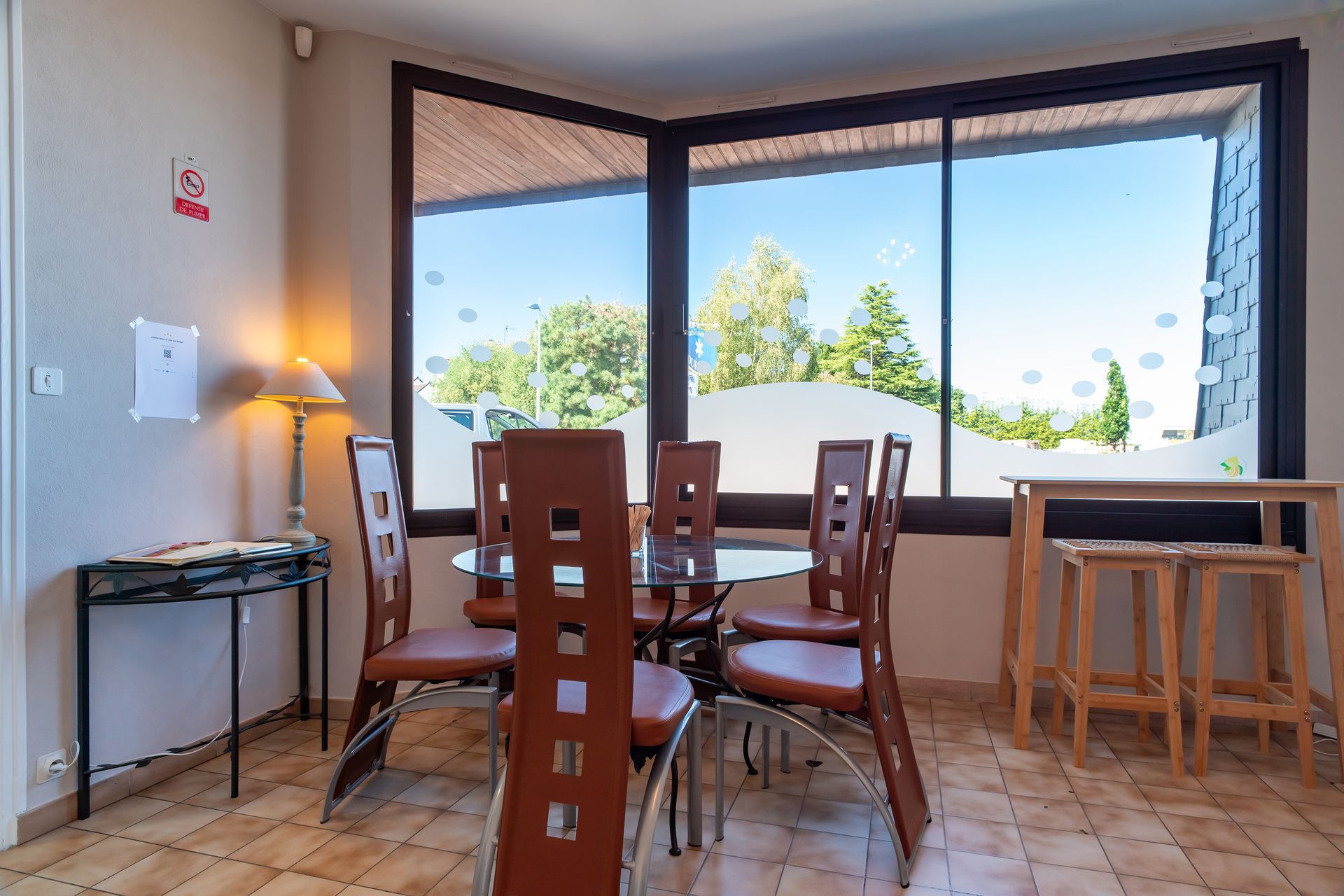 Coin de recueil avec une table ronde, des chaises marron et une grande fenêtre donnant sur les arbres.