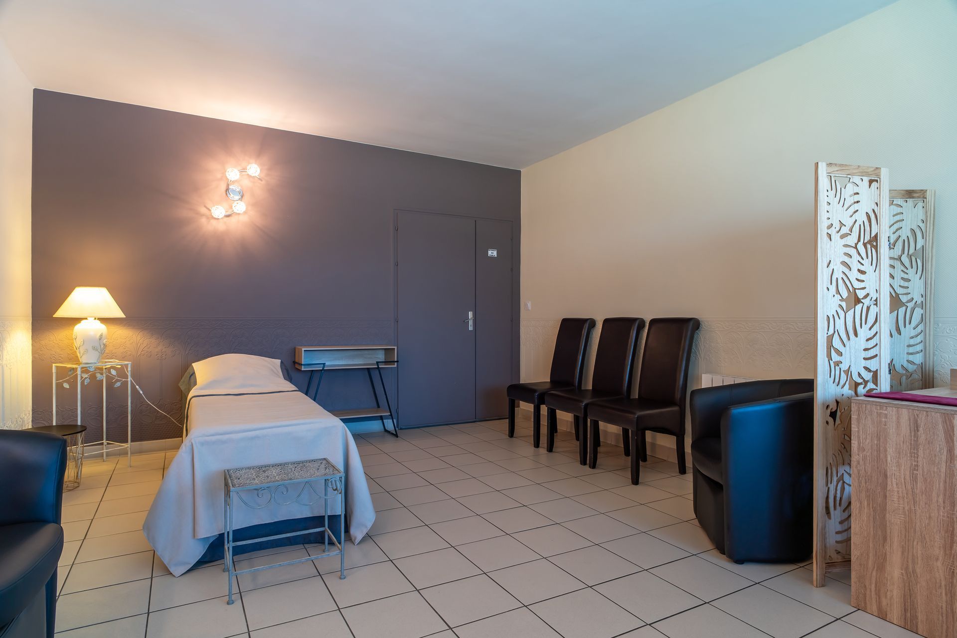 Salle de massothérapie avec lit de traitement, chaises et paravent décoratif.