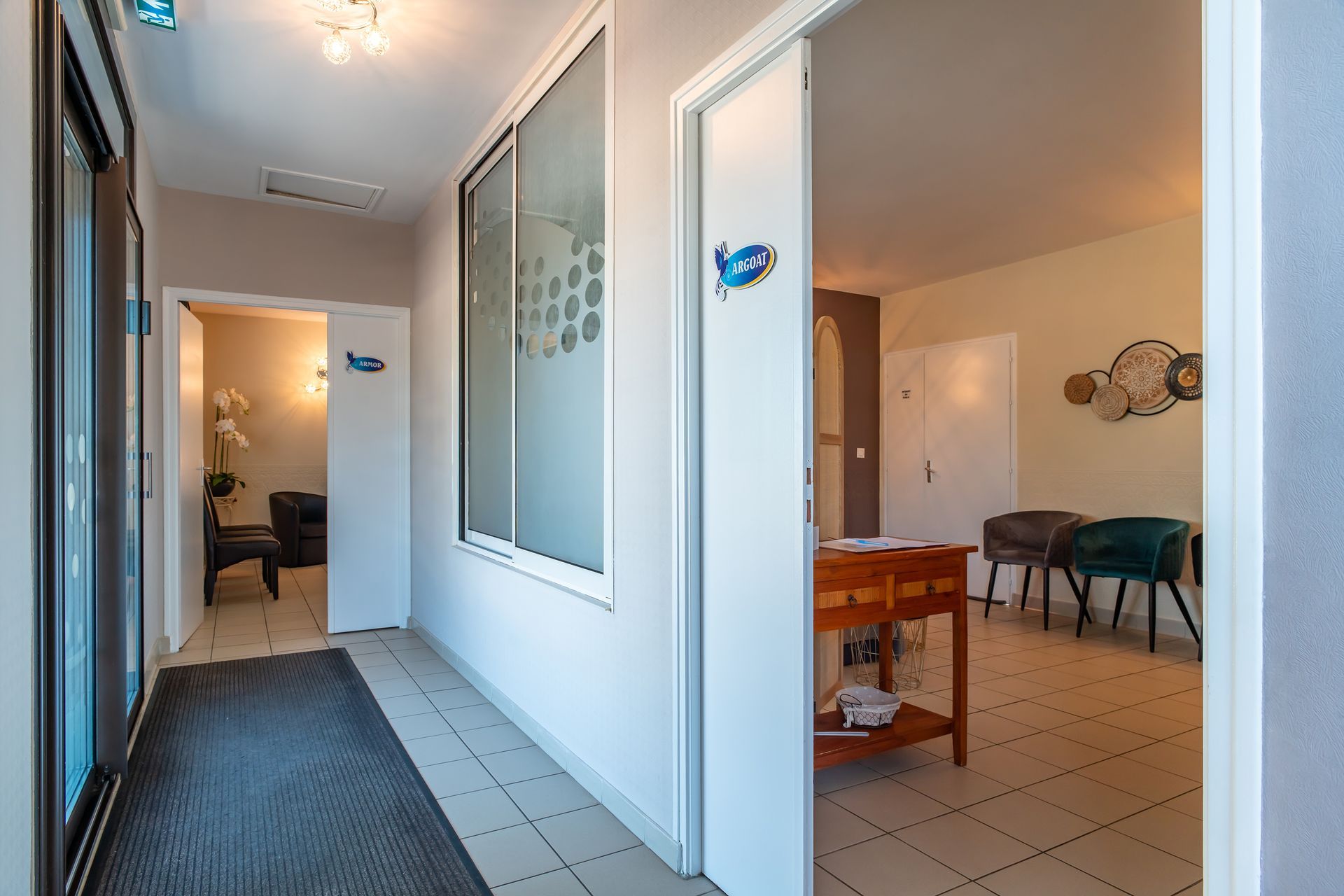 Couloir avec portes, fenêtre et salle d'attente. Murs clairs, tapis foncé. Touches bleues sur les portes.