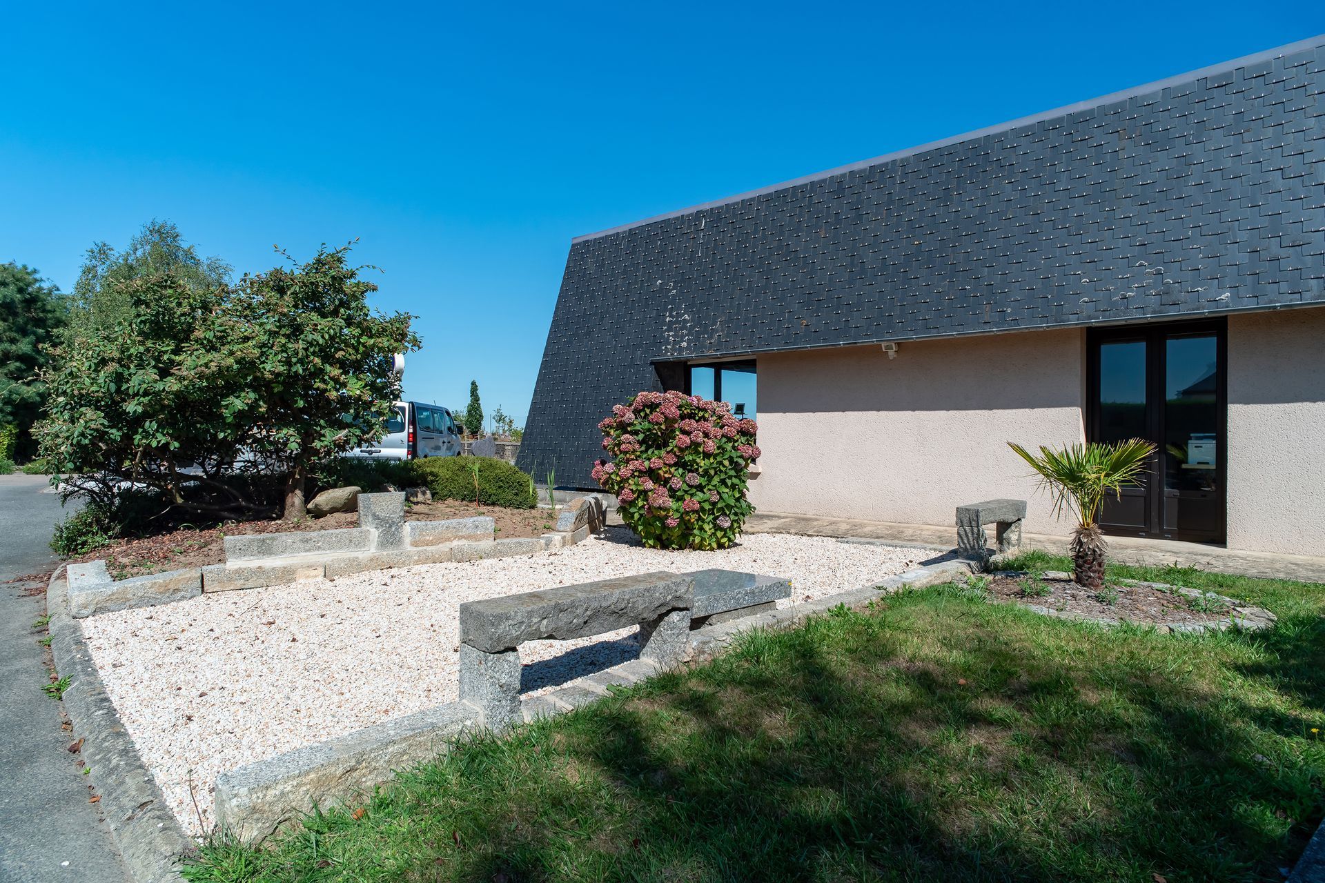 Extérieur d'un bâtiment avec toit en pente, bancs en pierre et aménagement paysager. Ciel bleu en arrière-plan.