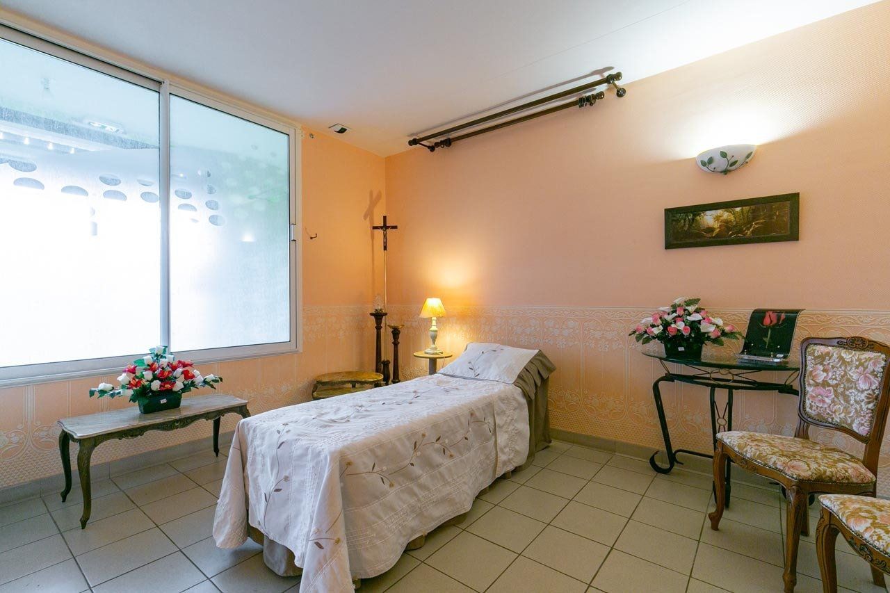 Une chambre avec un lit, deux chaises, une fenêtre et une table fleurie. Murs pastel et sol carrelé.