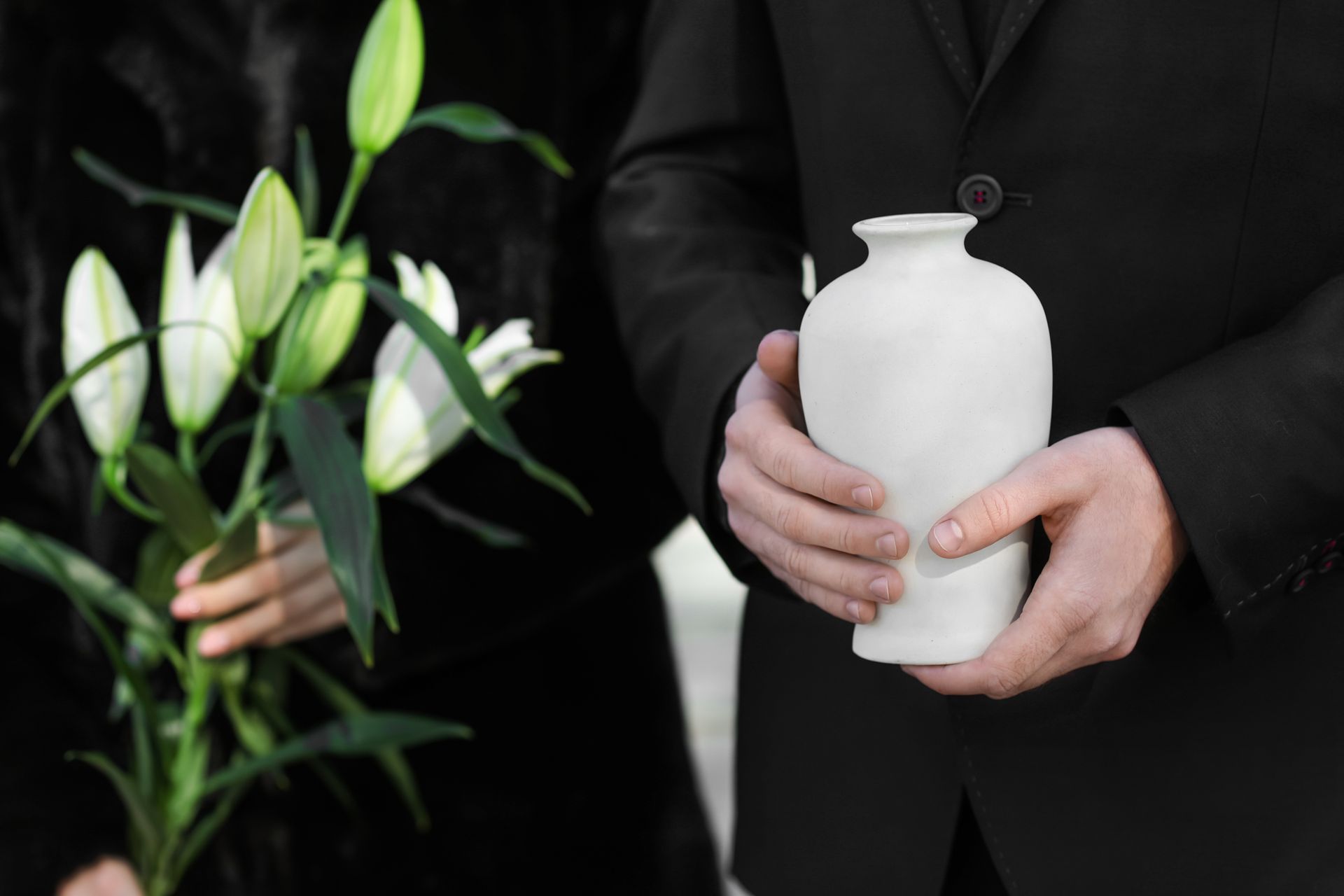 Une personne en costume noir tient une urne blanche, une autre personne tient des lys.
