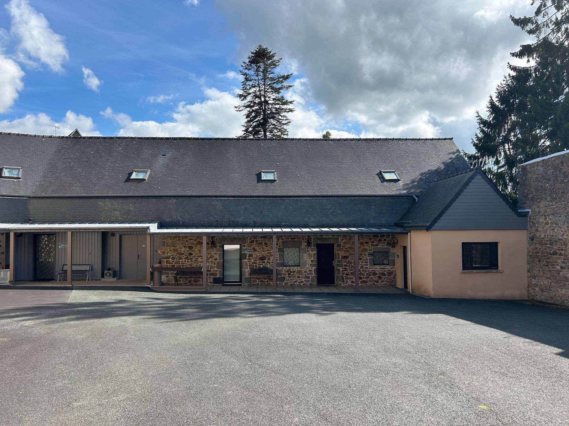 Bâtiment avec toit gris, alcôve remplie de bois et parking en gravier sous un ciel bleu.