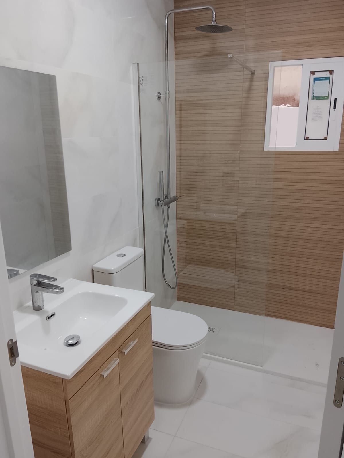 Baño moderno con lavabo, inodoro y ducha blancos; mueble de lavabo de madera y pared de ducha alicatada.