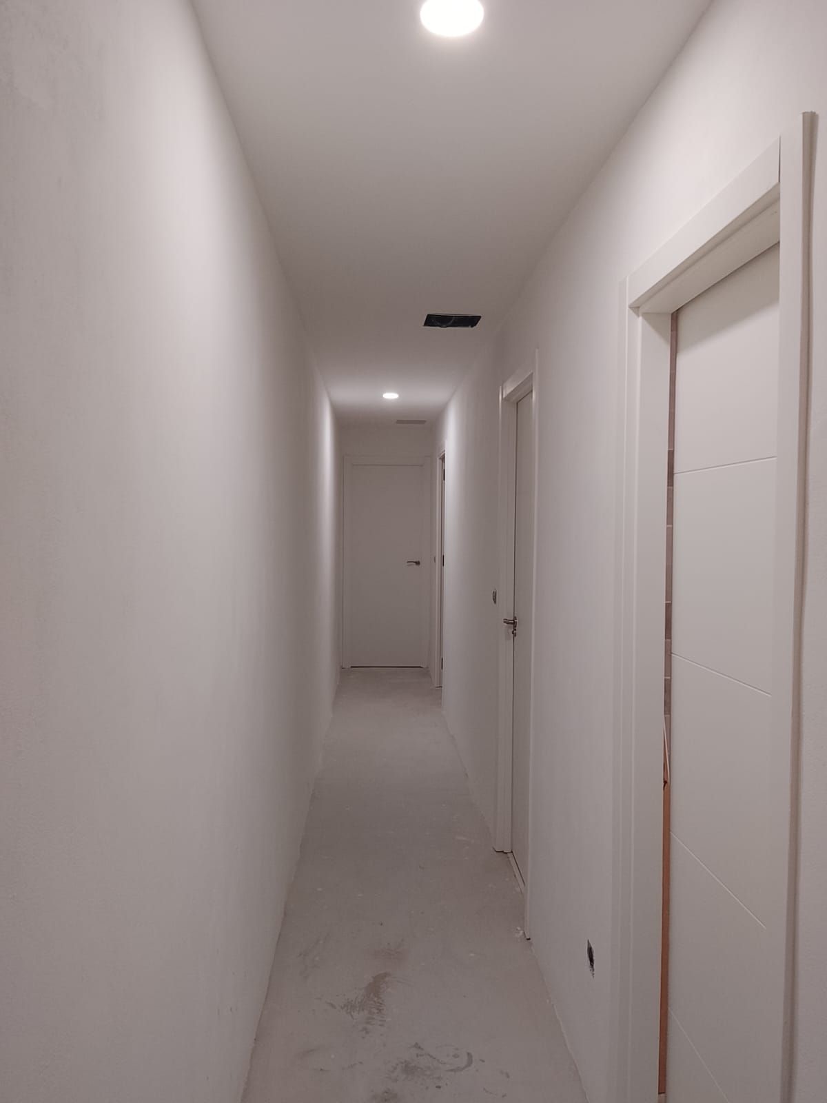 Pasillo estrecho y blanco con puertas, luz en el techo y suelo sin terminar.