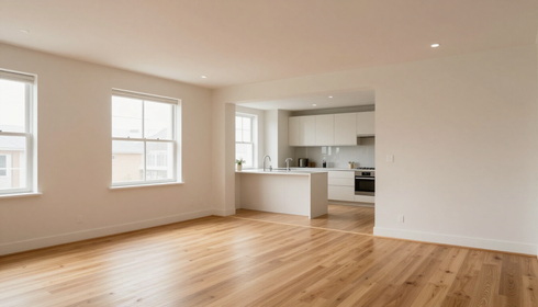 Sala de estar amplia y despejada, con suelo de madera, cocina abierta, paredes blancas y dos ventanas.