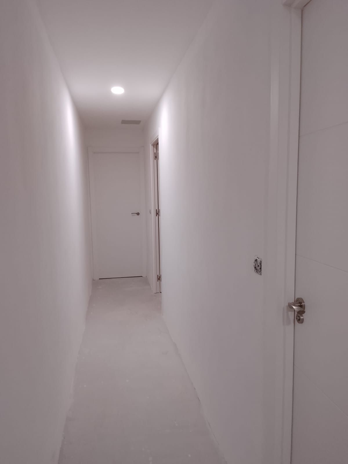 Pasillo estrecho y blanco con puertas y una luz en el techo, visto desde un extremo.