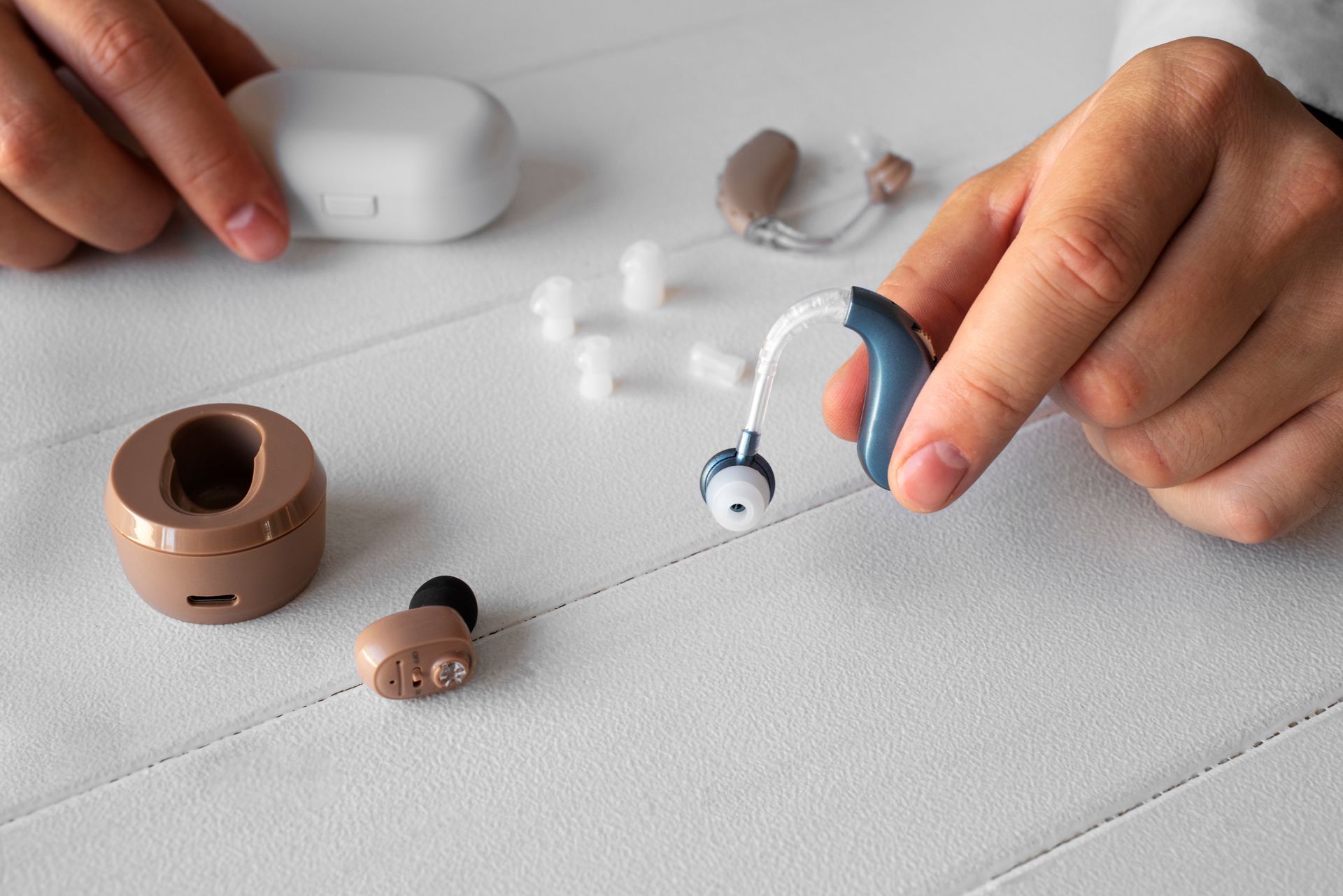 Persona sosteniendo un audífono, otros audífonos, un cargador y accesorios sobre una superficie blanca.