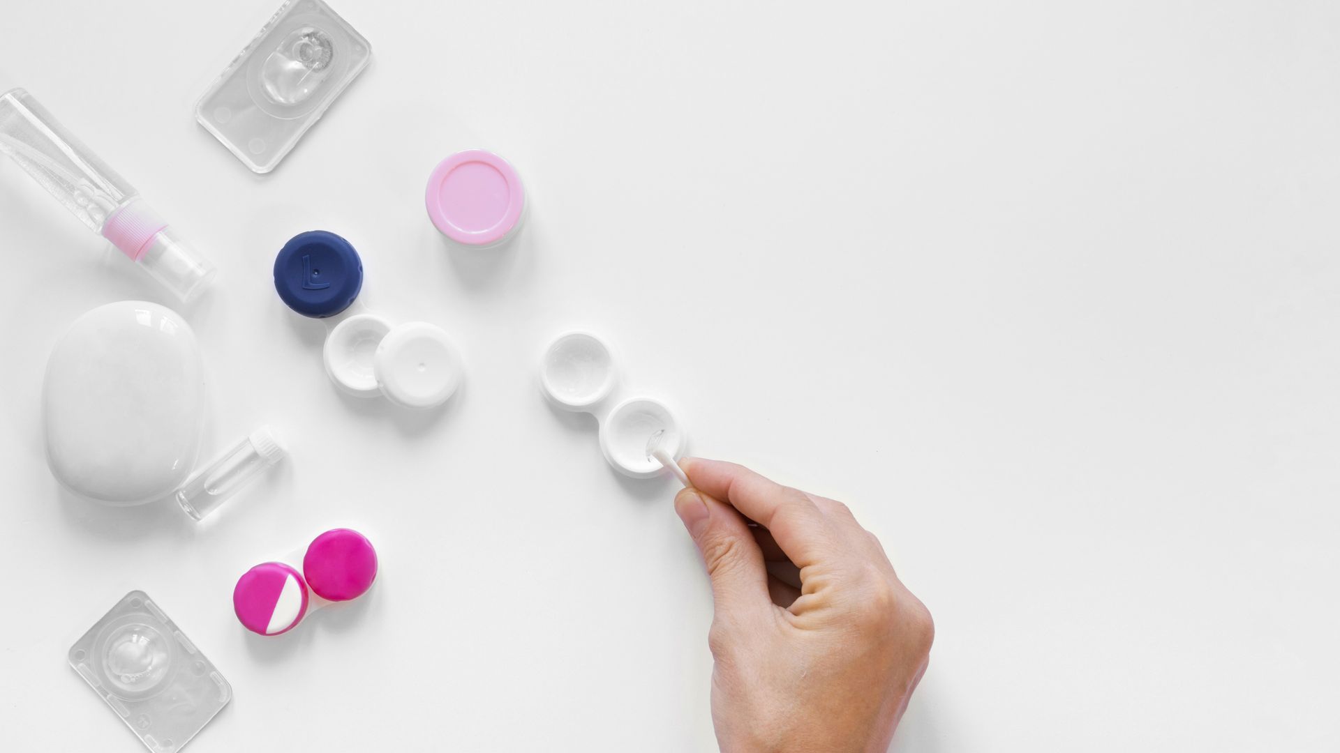 Mano que busca una lente de contacto, rodeada de estuches para lentes, solución y embalaje sobre una superficie blanca.