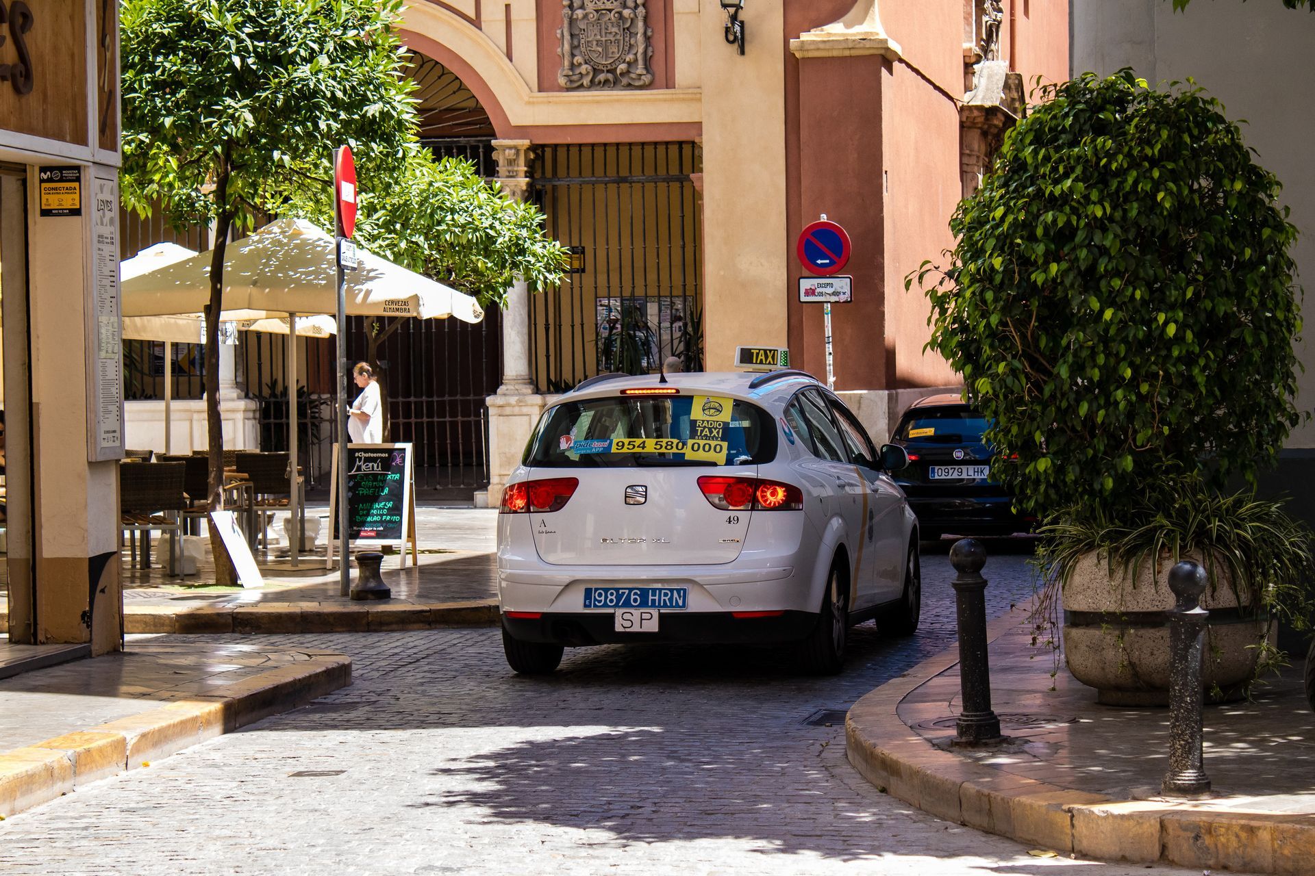 Un taxi blanco circula por una calle adoquinada en una ciudad europea, cerca de una cafetería.