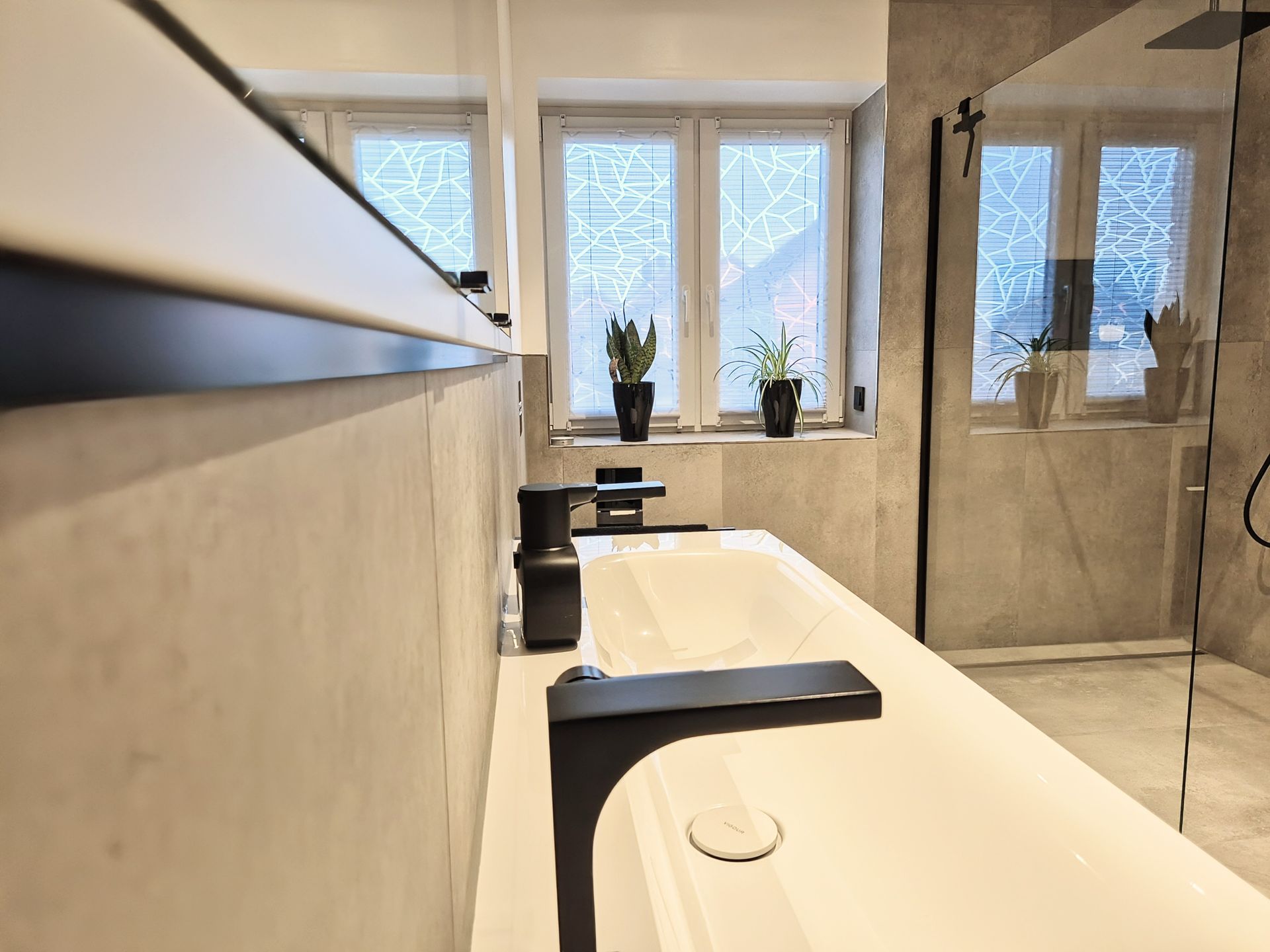 Seitlicher Blick auf ein weißes Waschbecken mit schwarzer Armatur in einem Badezimmer mit grauen Fliesen; im Hintergrund ein Fenster mit Pflanzen auf der Fensterbank und Sicht auf die bodengleiche Dusche.