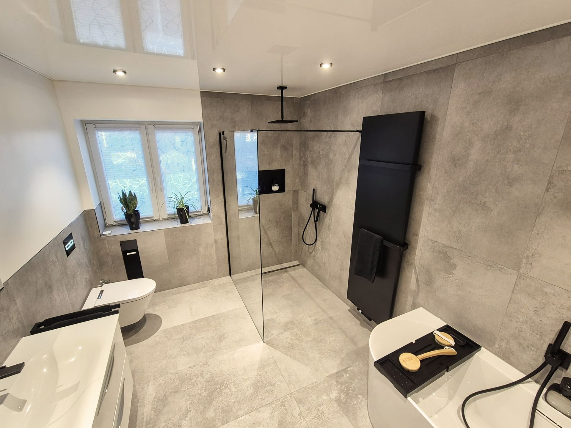 Weitwinkelaufnahme eines modernen Badezimmers mit freistehender Badewanne, bodengleicher Dusche, wandhängendem WC, Doppelwaschtisch und schwarzen Akzenten, inklusive Pflanzen auf der Fensterbank.