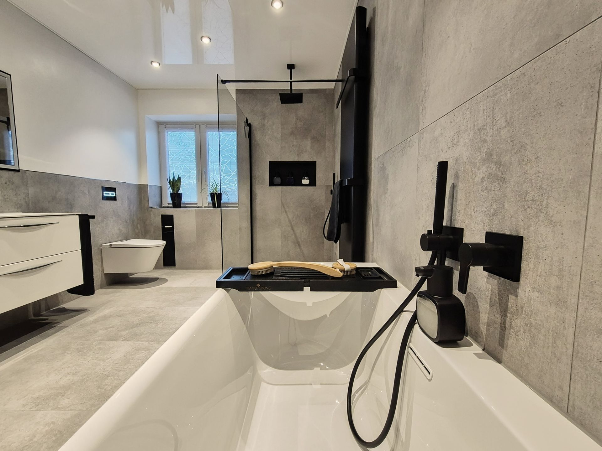 Perspektive aus der Badewanne auf das Badezimmer; sichtbar sind die schwarze Badewannenarmatur, die Dusche mit Glastrennwand sowie der Waschbereich und das wandhängende WC im Hintergrund.