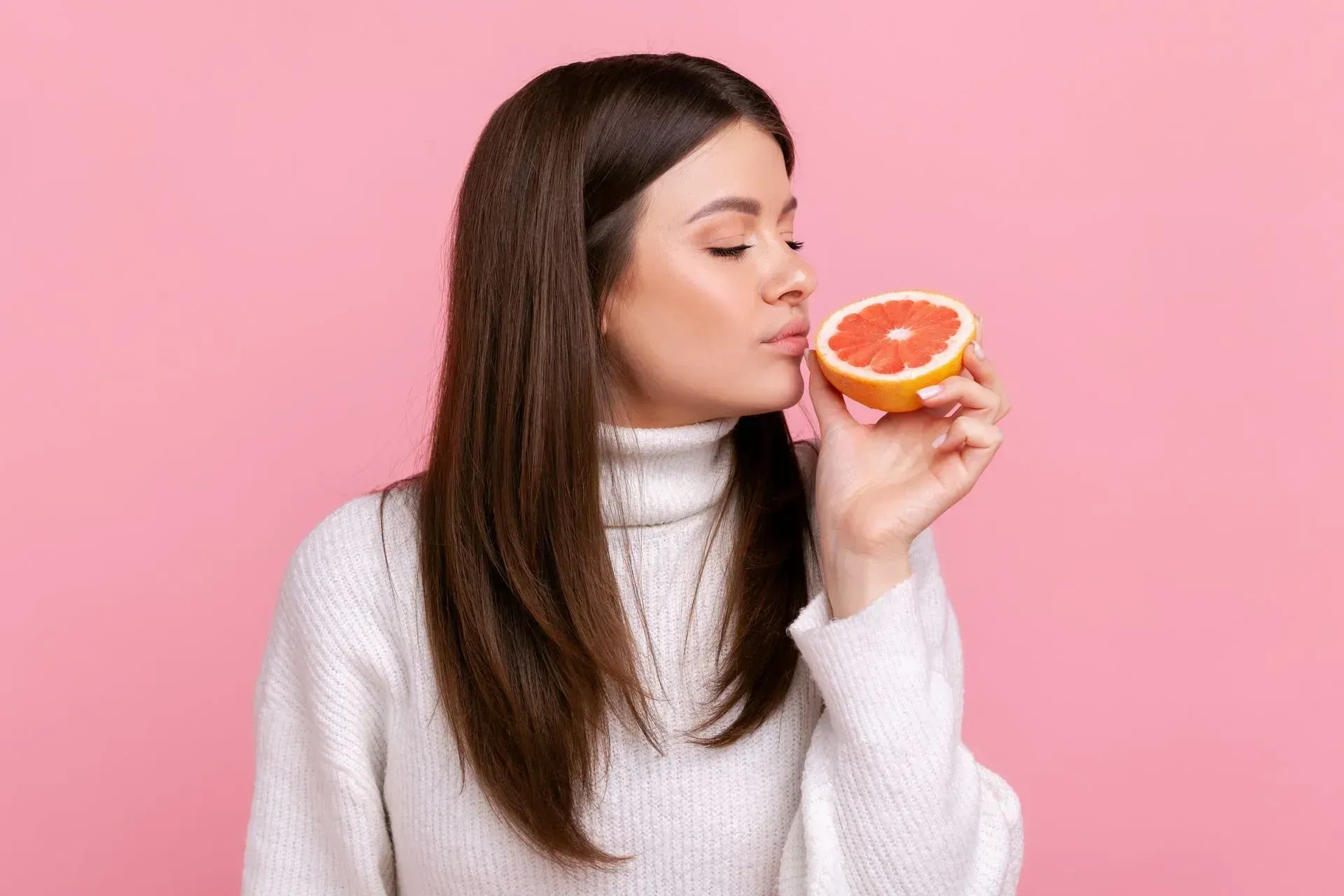 Mujer con un suéter blanco que sostiene y huele un pomelo sobre un fondo rosa