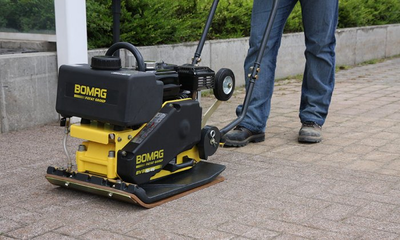 Ein Mann benutzt eine Bomag-Verdichtungsmaschine auf einem gepflasterten Gehweg.
