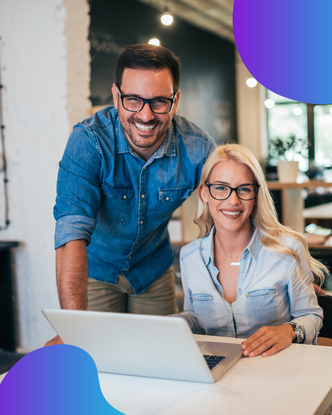 Un hombre y una mujer sonríen frente a una laptop en una oficina. Ambos llevan gafas y camisas vaqueras.