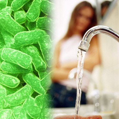 Bacterias verdes junto al agua corriente del grifo, con una mujer al fondo cerca de un lavabo.