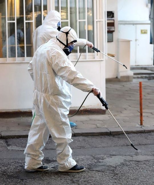 Dos personas con trajes de protección blancos rocían una sustancia en una acera cerca de la entrada de un edificio.