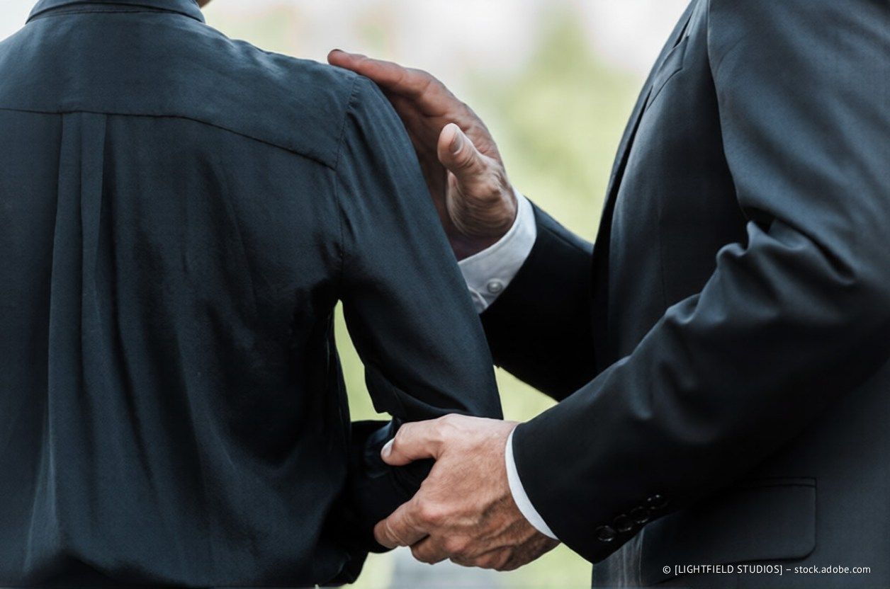 Eine Person im Anzug berührt die Schulter und den Arm einer anderen Person, möglicherweise um Trost oder Unterstützung anzubieten.