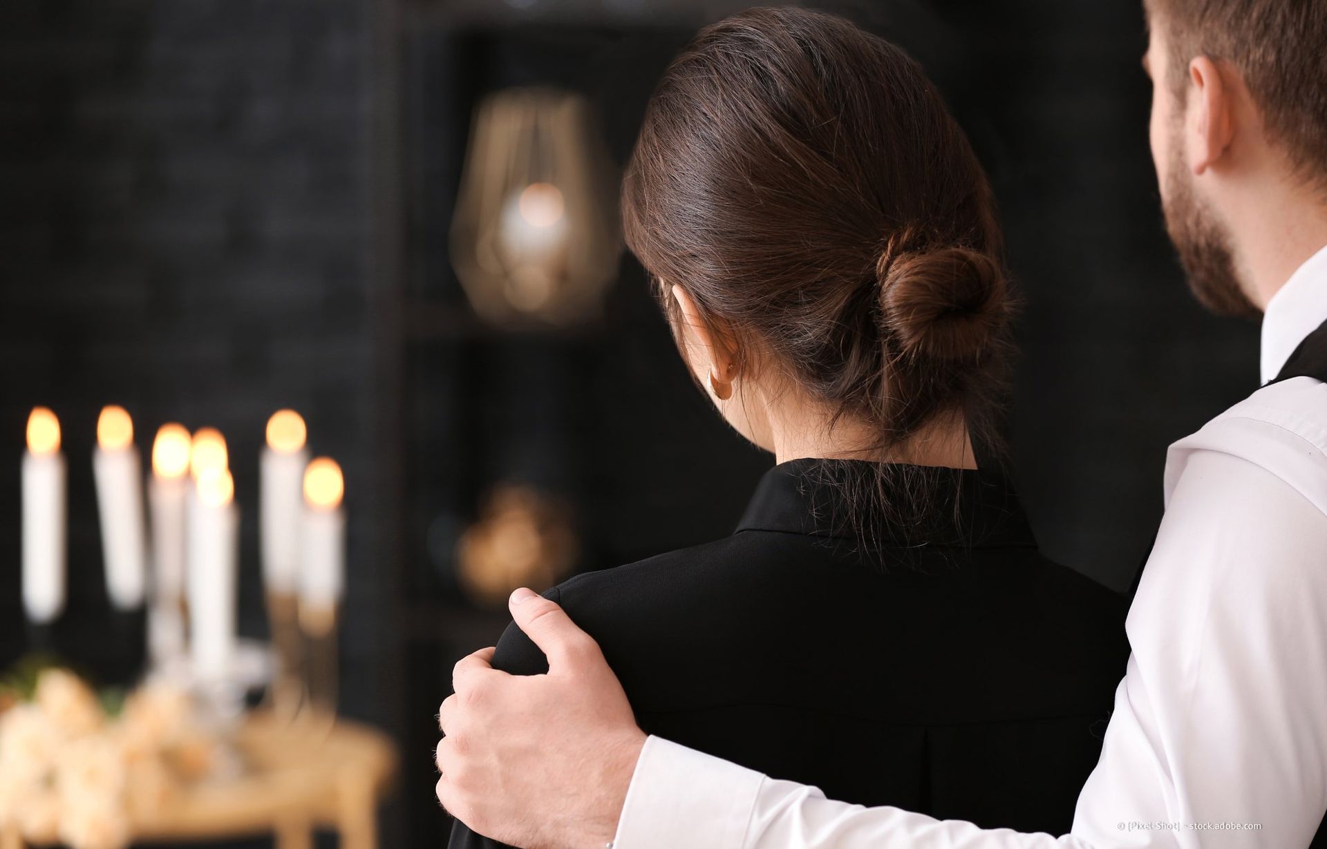 Eine Frau in Schwarz findet Trost in einer Hand auf ihrer Schulter, Kerzenlicht und einem verschwommenen Hintergrund.