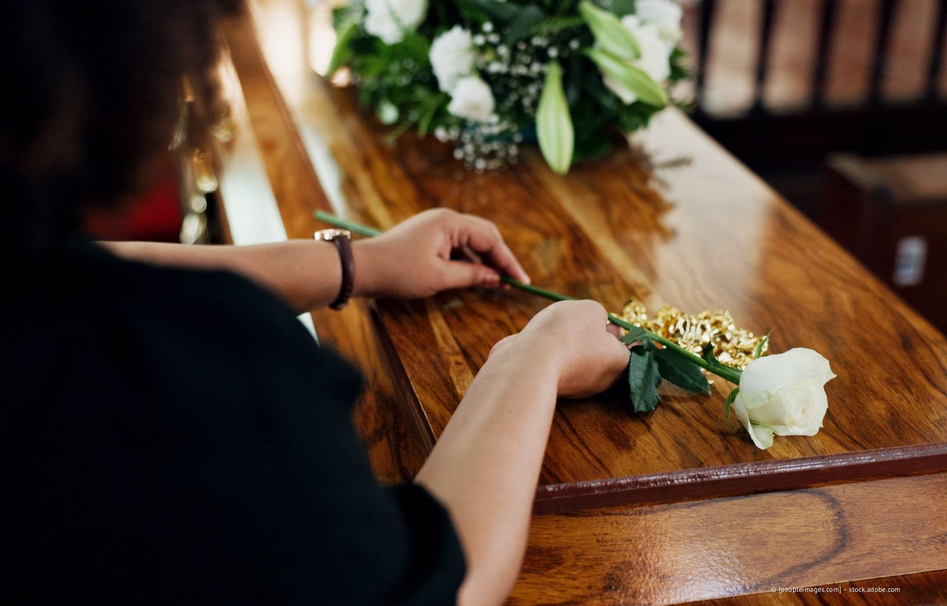 Eine Person legt eine weiße Rose auf einen geschlossenen Holzsarg, auf dem sich ein Blumenarrangement befindet.