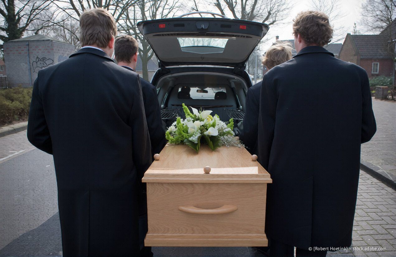 Vier Sargträger tragen einen Holzsarg zu einem offenen Kofferraum; darauf liegt ein Blumengesteck.
