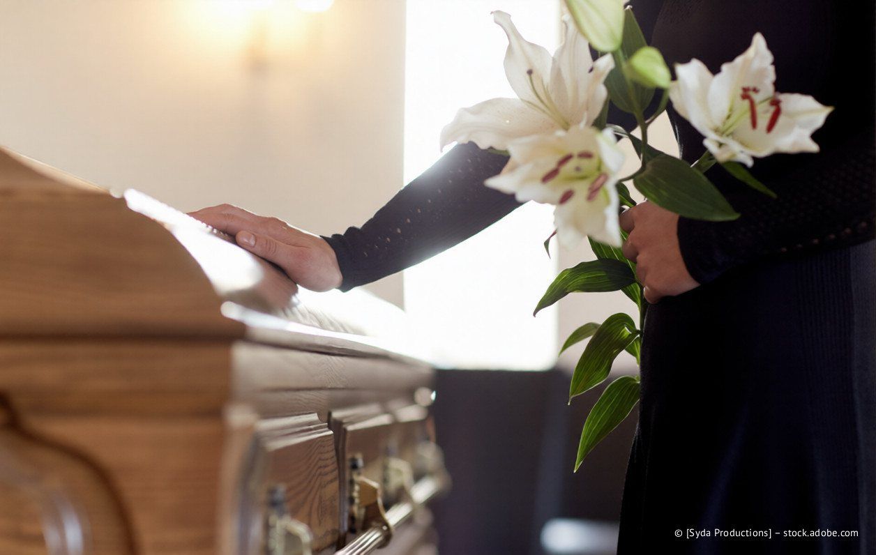 Eine Person hält weiße Lilien, die Hand liegt bei einer Beerdigung auf dem Sarg.