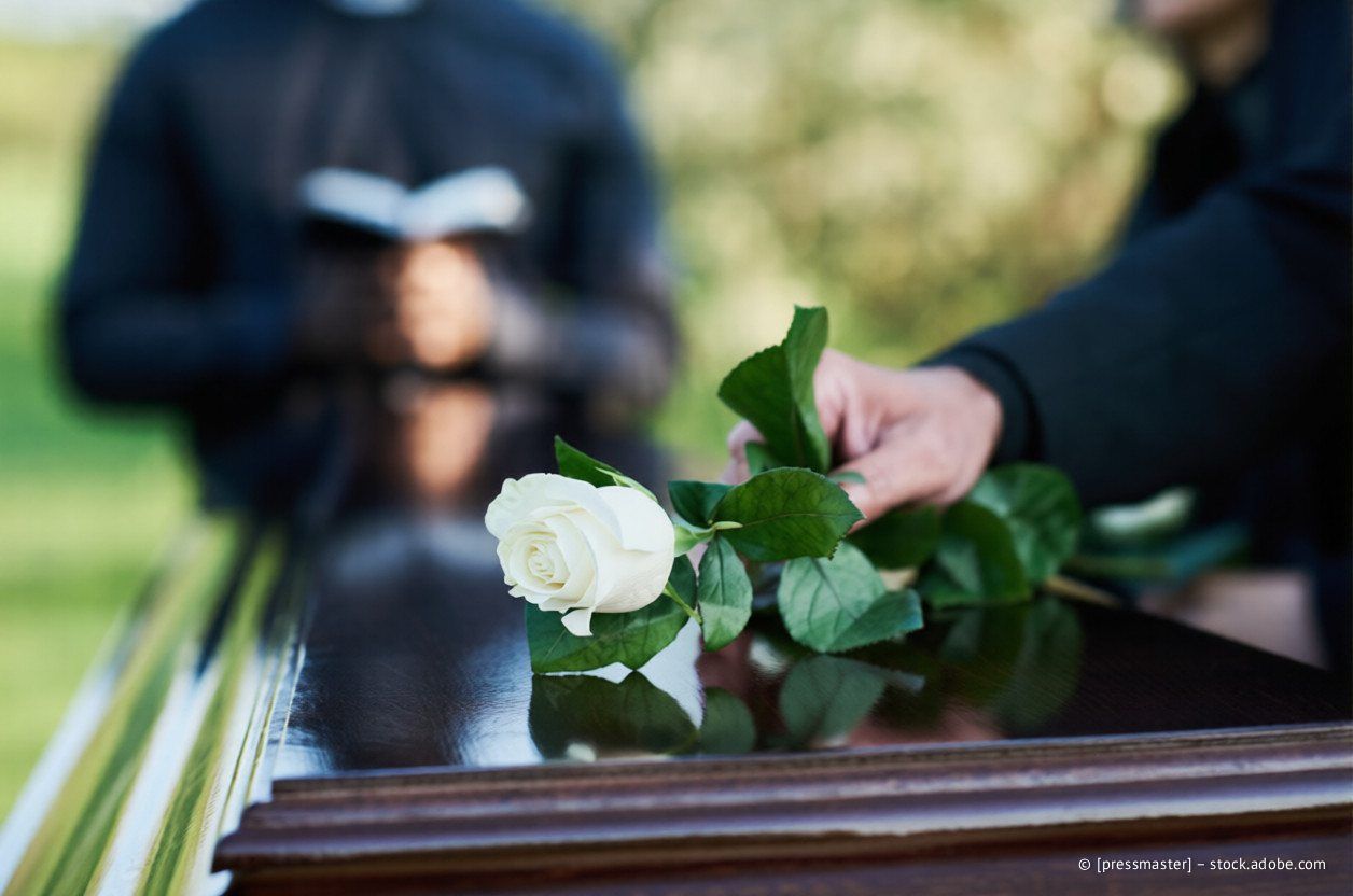 Eine Hand legt während einer Trauerfeier eine weiße Rose auf einen dunklen Holzsarg. Im Hintergrund liest eine verschwommene Gestalt.