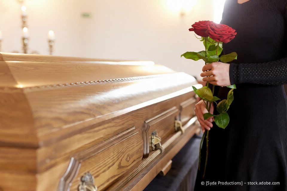 Eine Frau in schwarzem Kleid hält rote Rosen und steht in einem schwach beleuchteten Saal neben einem geschlossenen Holzsarg.