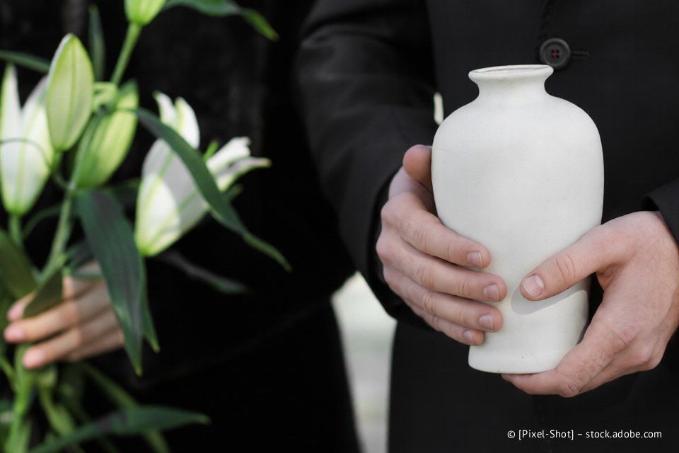 Eine Person hält eine weiße Urne mit Blumen und eine Person in schwarzer Kleidung.