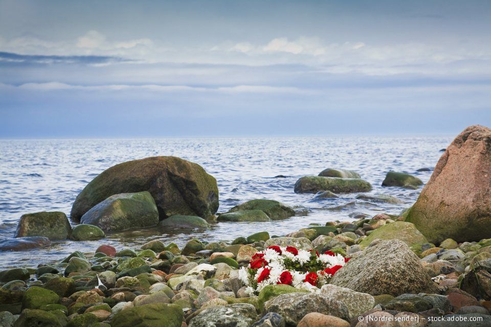 Ein Kranz aus roten und weißen Blumen liegt auf einem felsigen Strand am Meer.