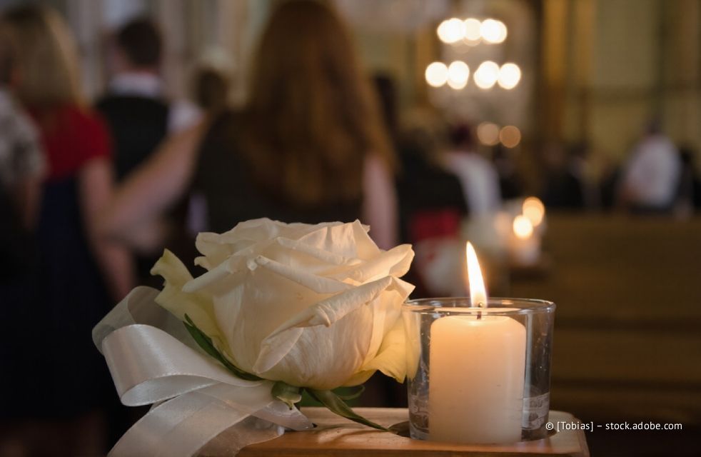 Weiße Rose und brennende Kerze auf einer Holzfläche, im Hintergrund verschwommene Personen und ein Kronleuchter.