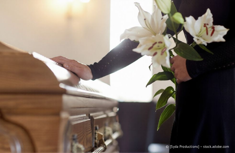 Eine Person in schwarzer Kleidung hält weiße Lilien und legt während der Beerdigung die Hand auf einen Holzsarg.
