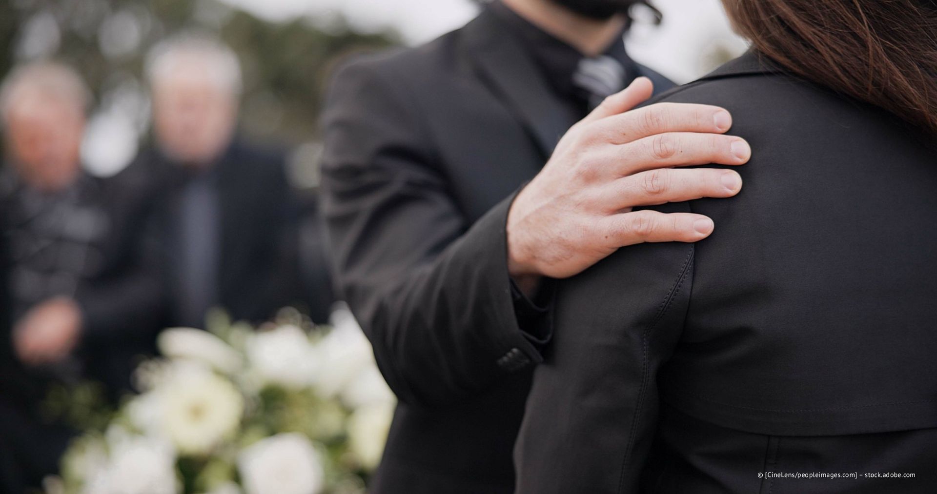 Eine Person tröstet eine andere bei einer Beerdigung; Hand auf der Schulter, schwarze Kleidung, weiße Blumen im Vordergrund, verschwommene Gestalten im Hintergrund.