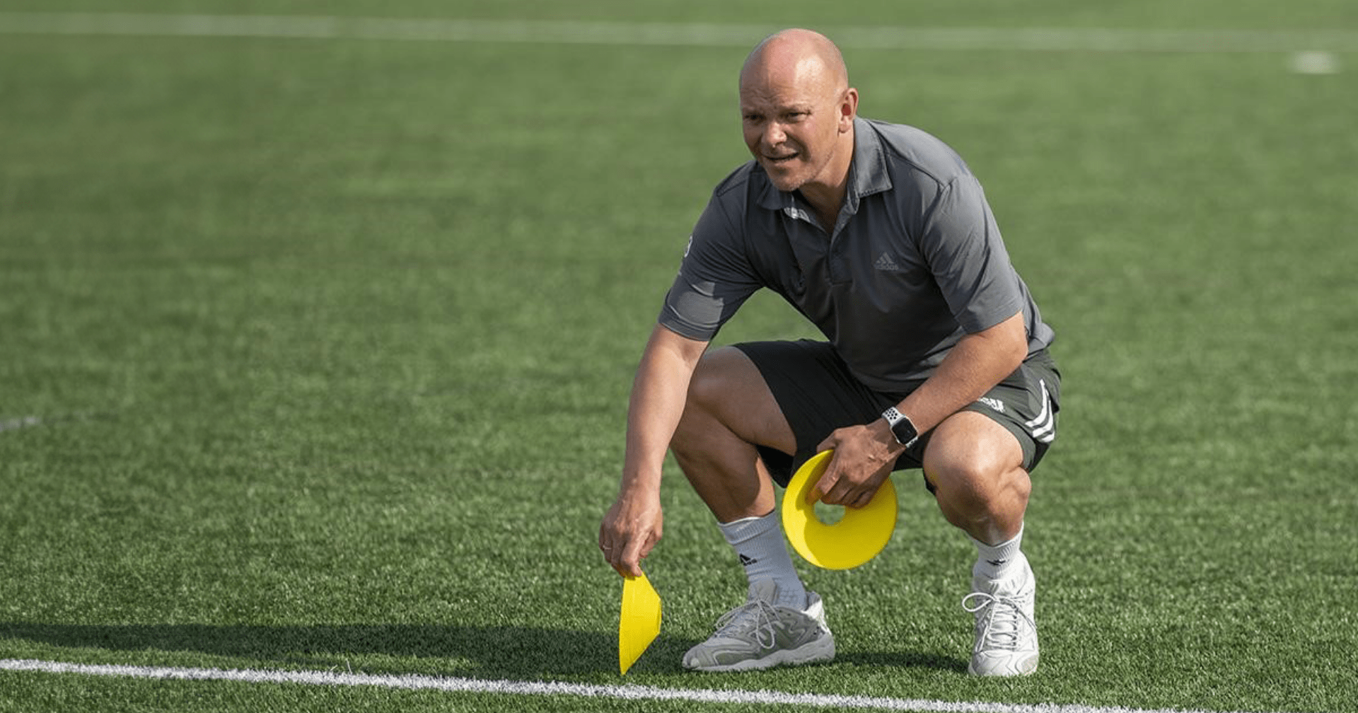 A man is squatting on a soccer field holding a yellow cone
